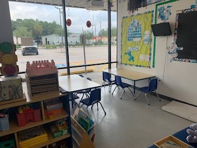 A brightly lit classroom interior with tables and chairs, a large window overlooking a parking lot, and shelves holding colorful learning materials.