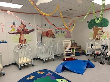 A brightly decorated nursery with cribs, a rocking chair, and blue play mats; decorated with Winnie the Pooh characters and colorful streamers.