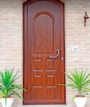 Porta d'ingresso in legno con maniglia in metallo, incorniciata da due piante verdi in vaso addossate a un muro di mattoni.