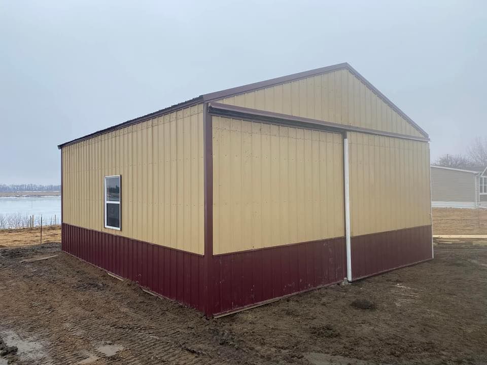 A tan and red metal building with a sliding door is sitting on top of a dirt field.