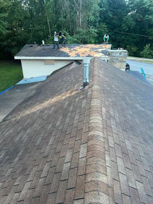 Two workers perform a roof replacement on a shingled roof, with one section stripped to the bare wood.