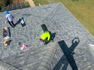 Two workers in safety gear installing grey shingles on a residential roof.