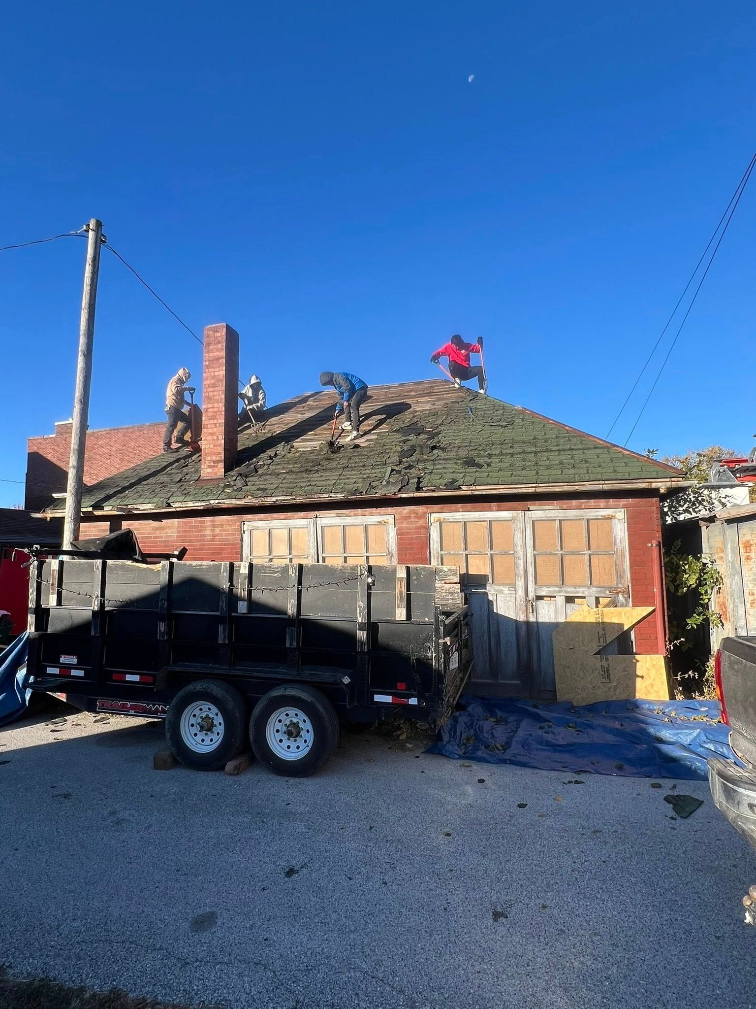 Construction workers repair the shingled roof of a brick building near a trailer under a clear blue sky.