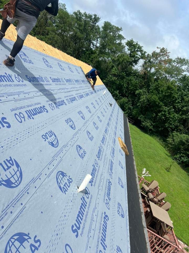Two workers installing blue Atlas Summit 60 underlayment on a wooden roof deck overlooking a grassy yard.