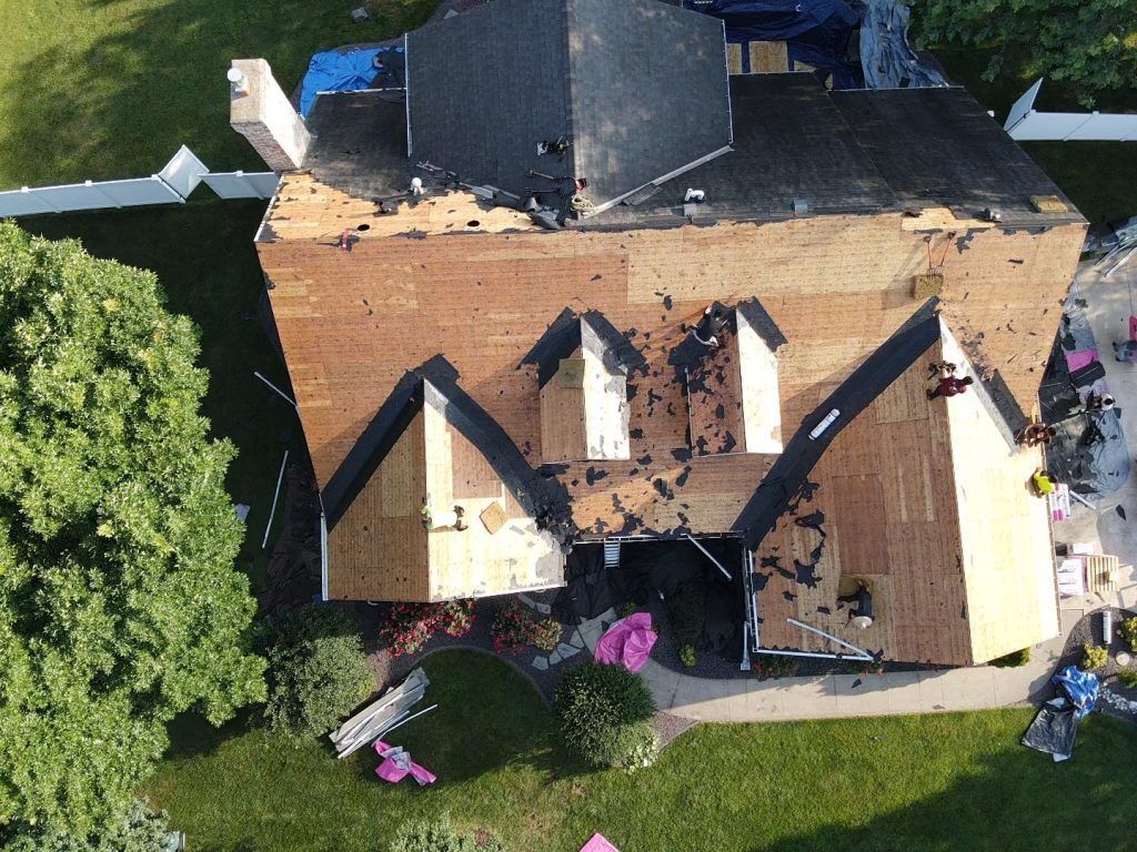 An aerial view shows a residential roof being replaced, with sections of shingles removed to expose the wooden deck.