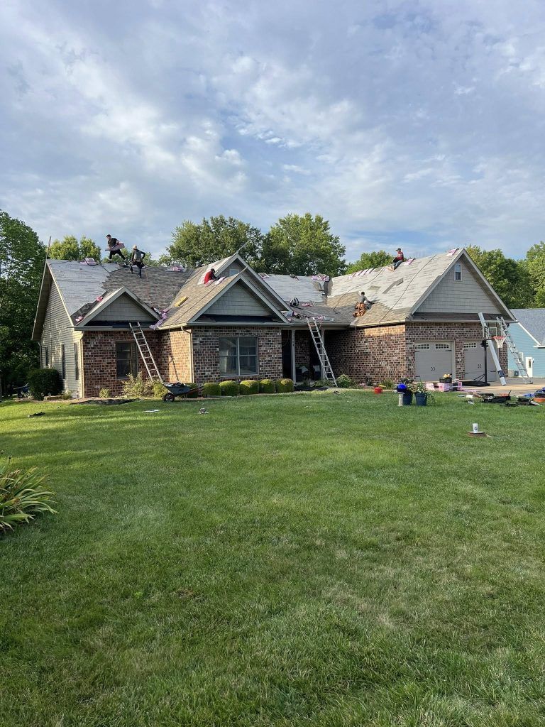 Workers perform roof repairs on a brick house during the day, with ladders positioned against the exterior walls.