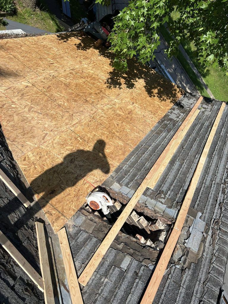 An overhead view of a roof under construction, showing plywood sheathing, exposed wooden rafters, and a chimney.