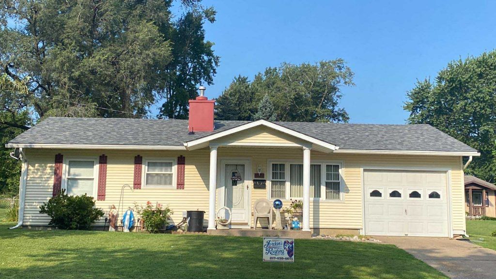 A single-story, light yellow house with a gray shingle roof, a red chimney, and a small front porch on a grassy lawn.