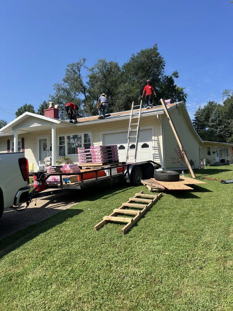 Three people work on a house roof with a trailer parked in the driveway and a ladder on the grass in front of it.