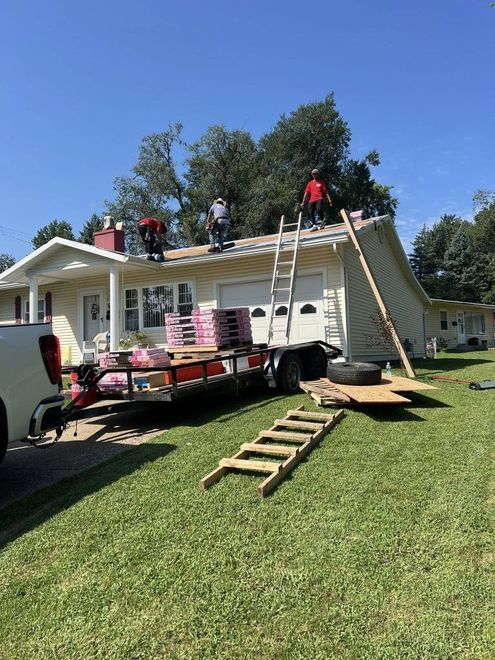 Workers in red shirts install a new roof on a light-colored house, using ladders and a trailer filled with shingles.