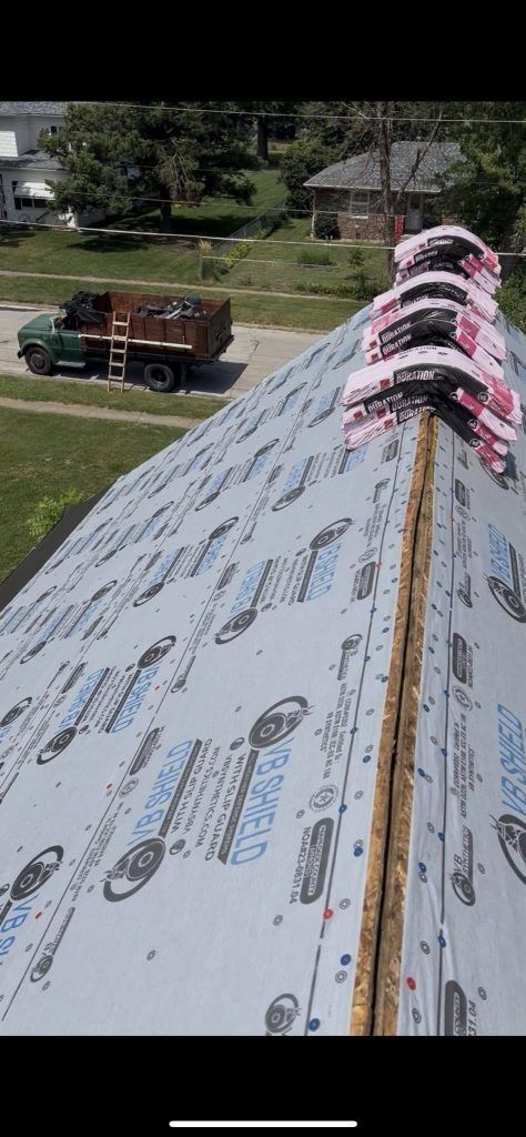 A residential roof covered in grey synthetic underlayment with stacks of shingle bundles placed along the ridge line.