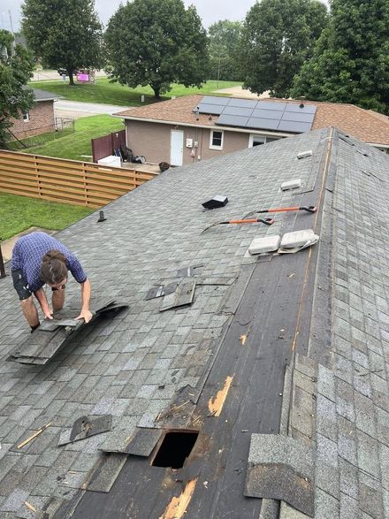 A person in a blue shirt repairs a gray shingle roof, working near an open hole in the roof deck.