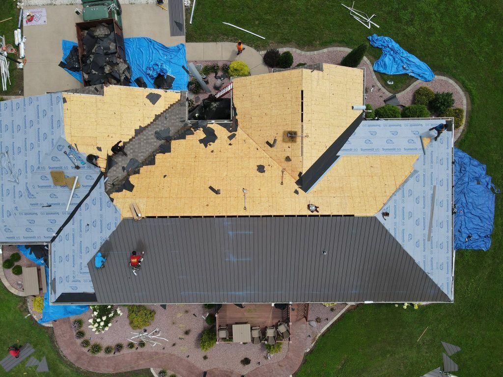 An aerial view shows a residential roof under construction, with sections featuring bare plywood, underlayment, and shingles.