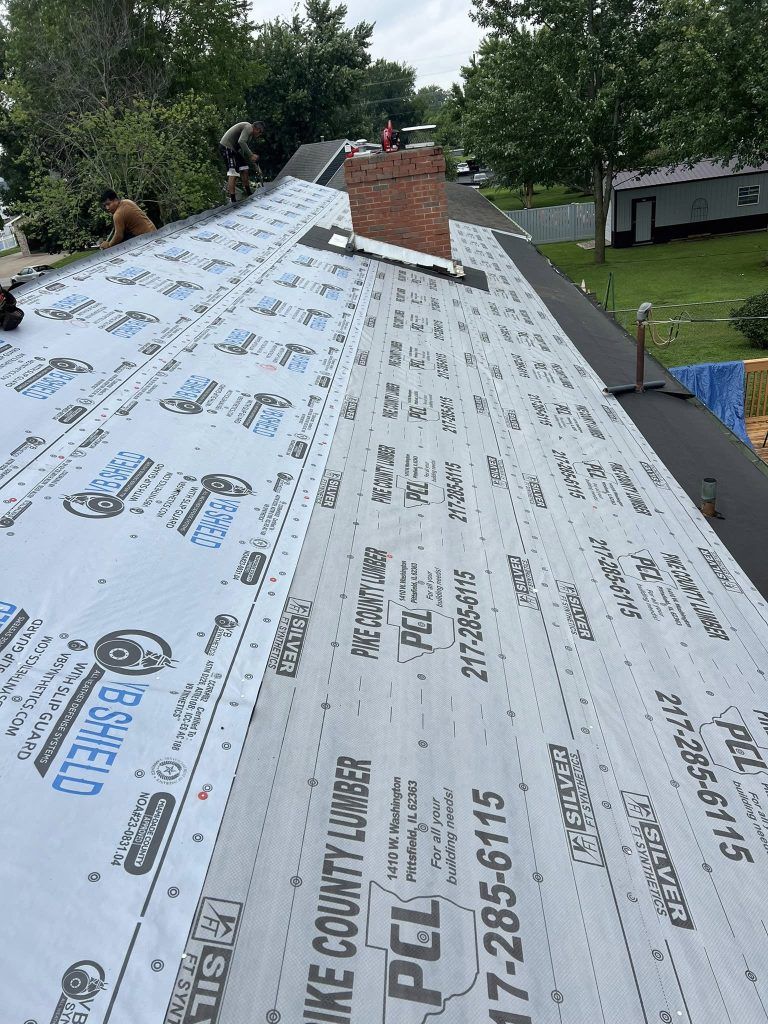 Roofers working on a residential roof covered in white underlayment, featuring a brick chimney, in a grassy backyard.