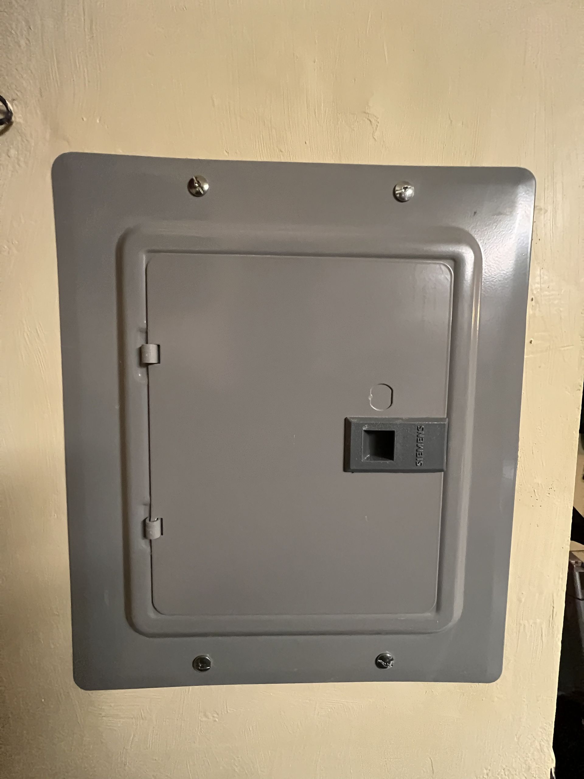 Gray electrical panel cover mounted on a beige wall with a small latch handle.