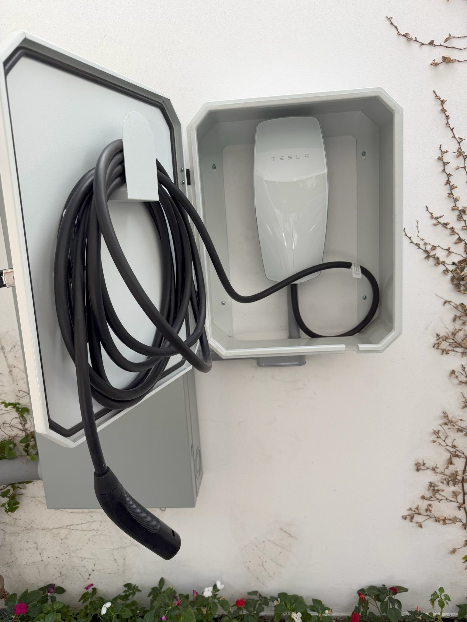 Open wall-mounted EV charging box with coiled black cable and white charger unit in a gray exterior cabinet.