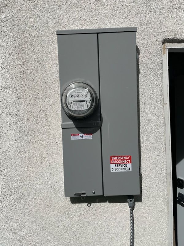Gray electrical meter box mounted on an exterior stucco wall with warning labels and conduit cable