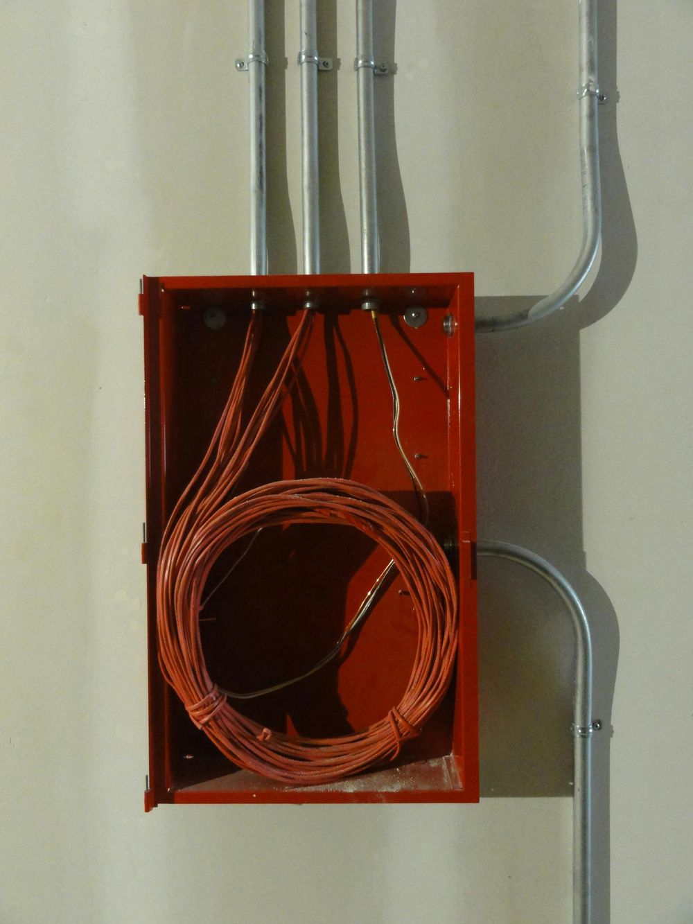 Open red electrical junction box with coiled copper wiring and gray conduit pipes on a wall