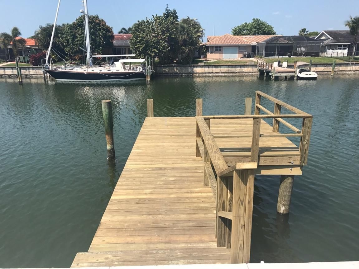 A wooden dock with a sailboat in the background with a residential home in the background.