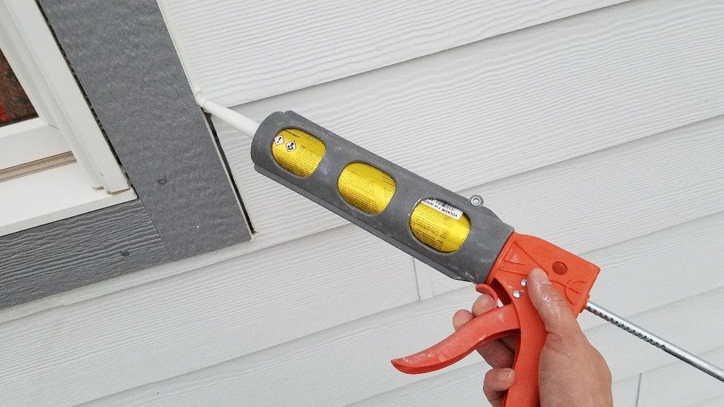 Person using a caulk gun to seal around a window frame on a white siding exterior.