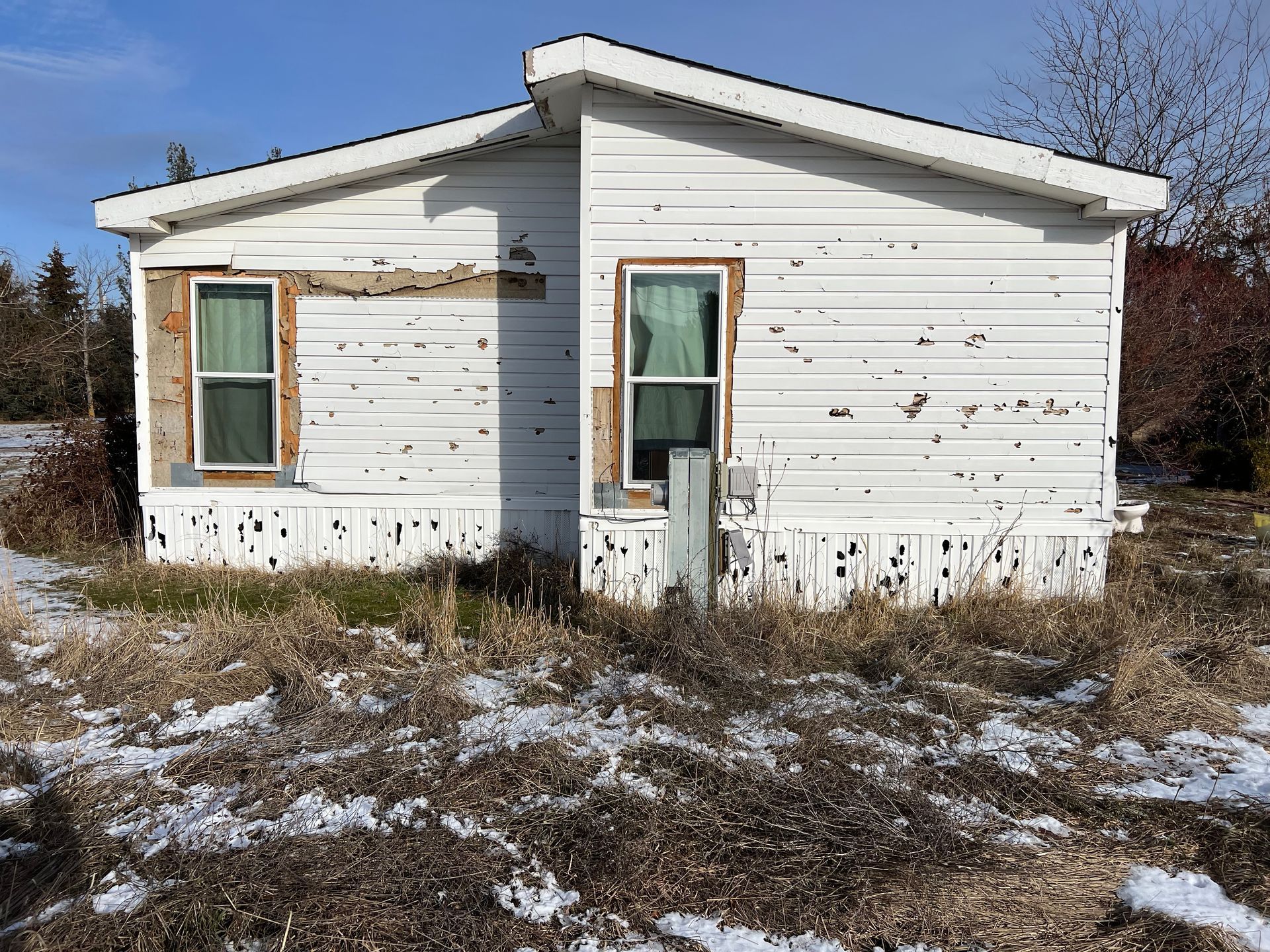 Dilapidated white mobile home with peeling siding, set in a field with patches of snow.