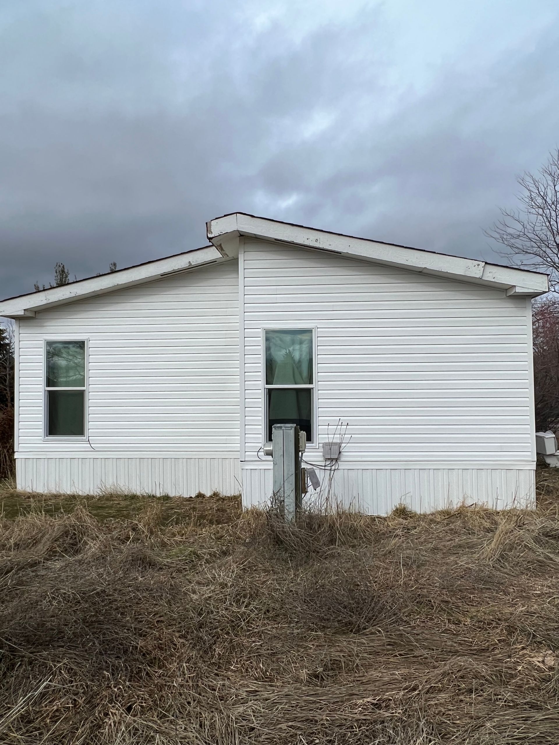 White mobile home with two windows under a cloudy sky, in front of overgrown, dry grass.