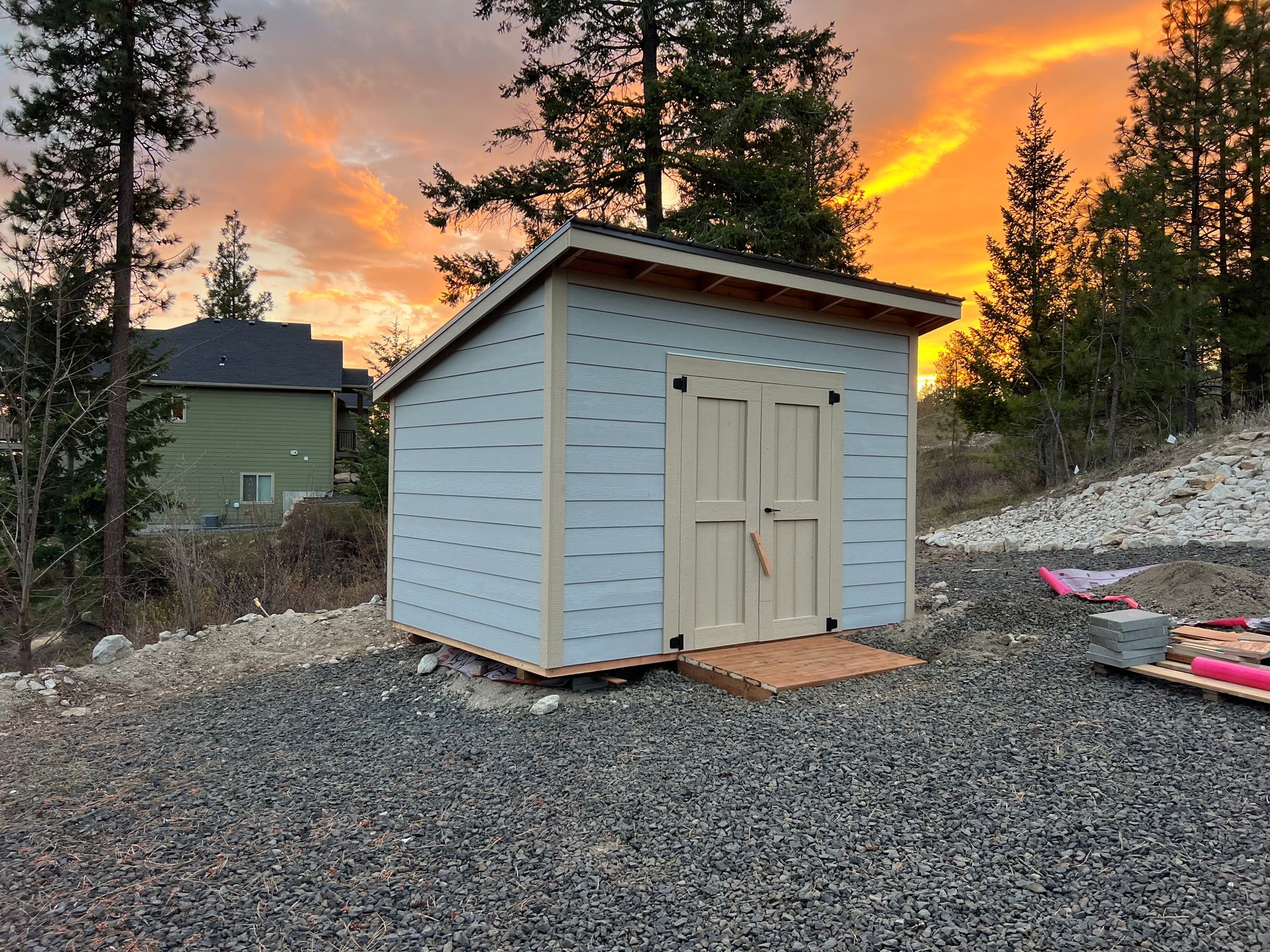 Blue shed with light brown doors and ramp; rocky ground, sunset sky.