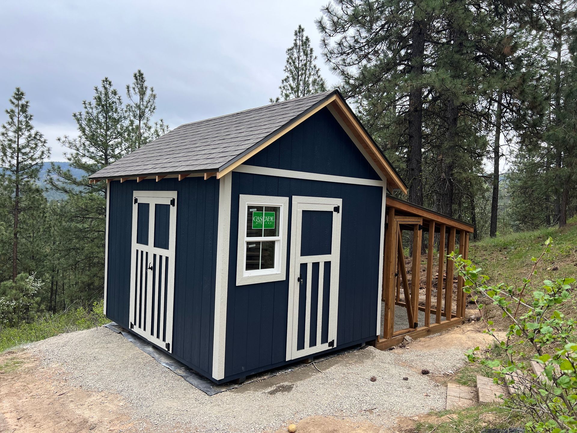 Blue shed with white trim and a brown wooden side structure, set in a wooded area.