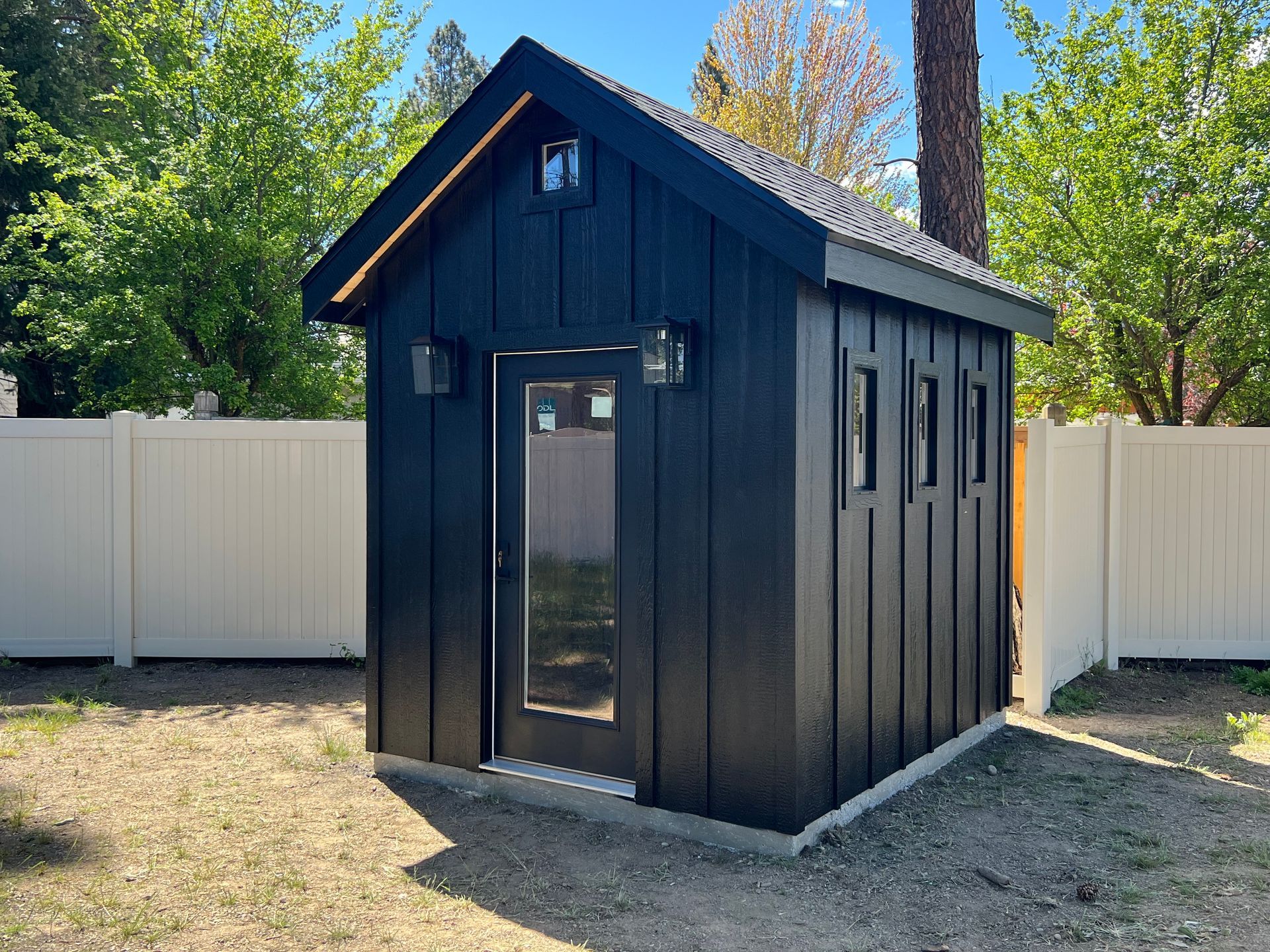 Black backyard shed with door, windows, and lights; set on a concrete foundation, next to a white fence.