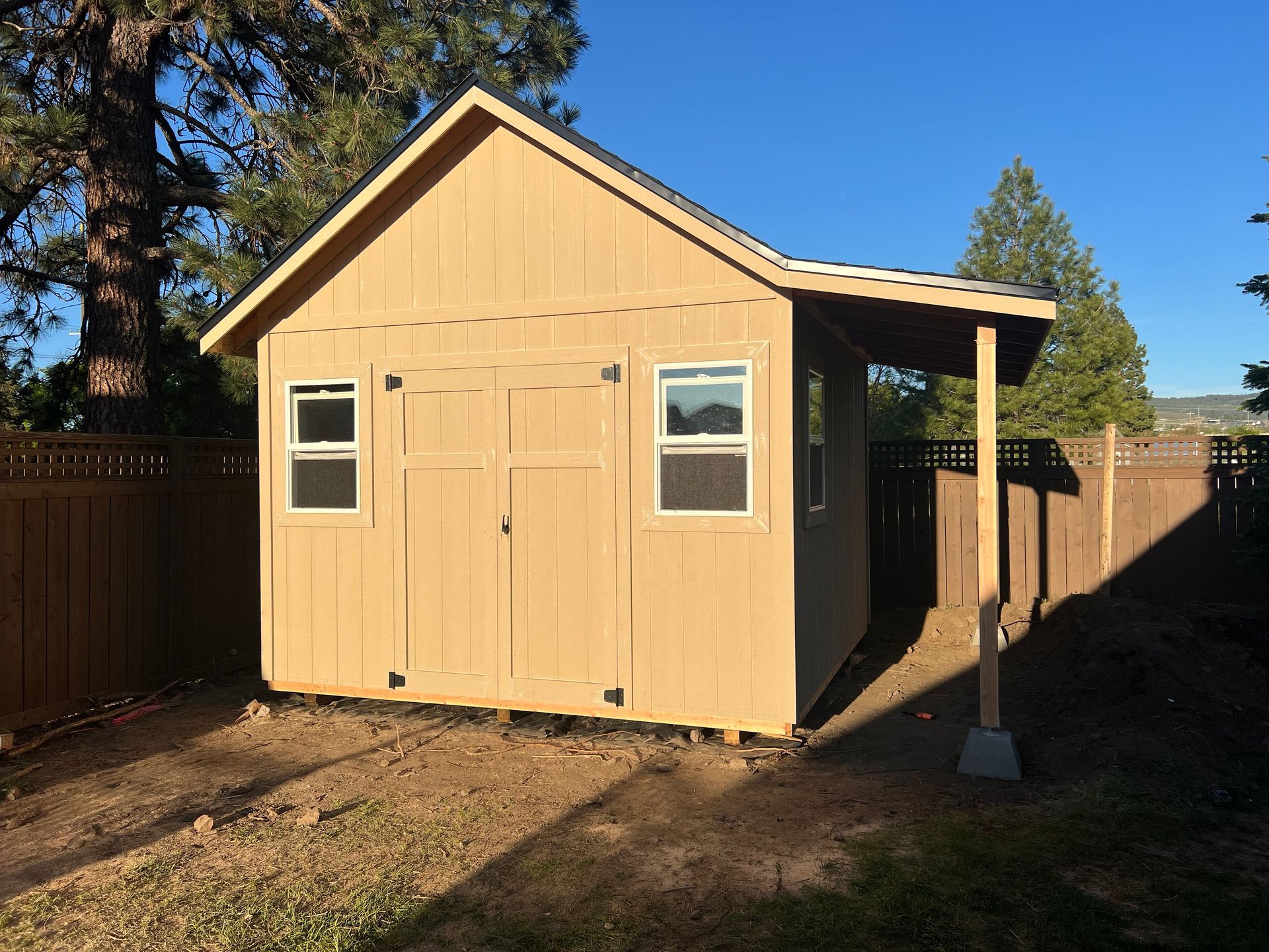 Tan shed with a gabled roof and small awning, set in a backyard with a wooden fence.