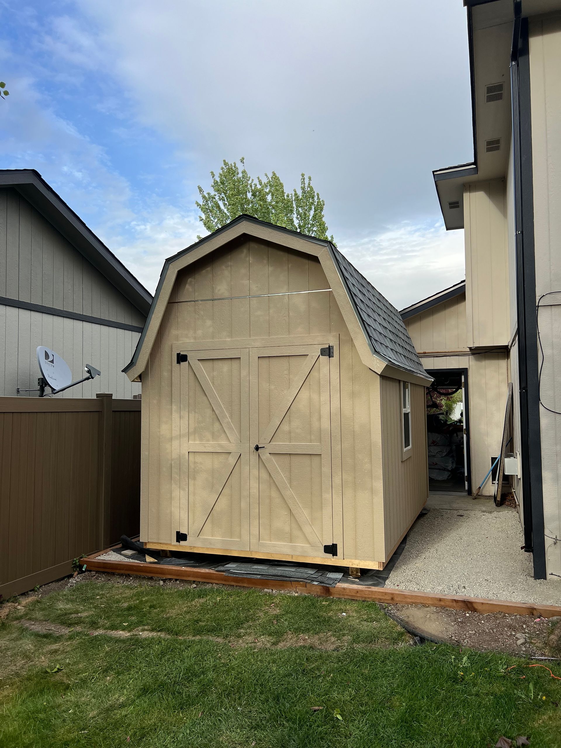 Wooden barn-style shed in a backyard, next to a house. Brown and tan colors.