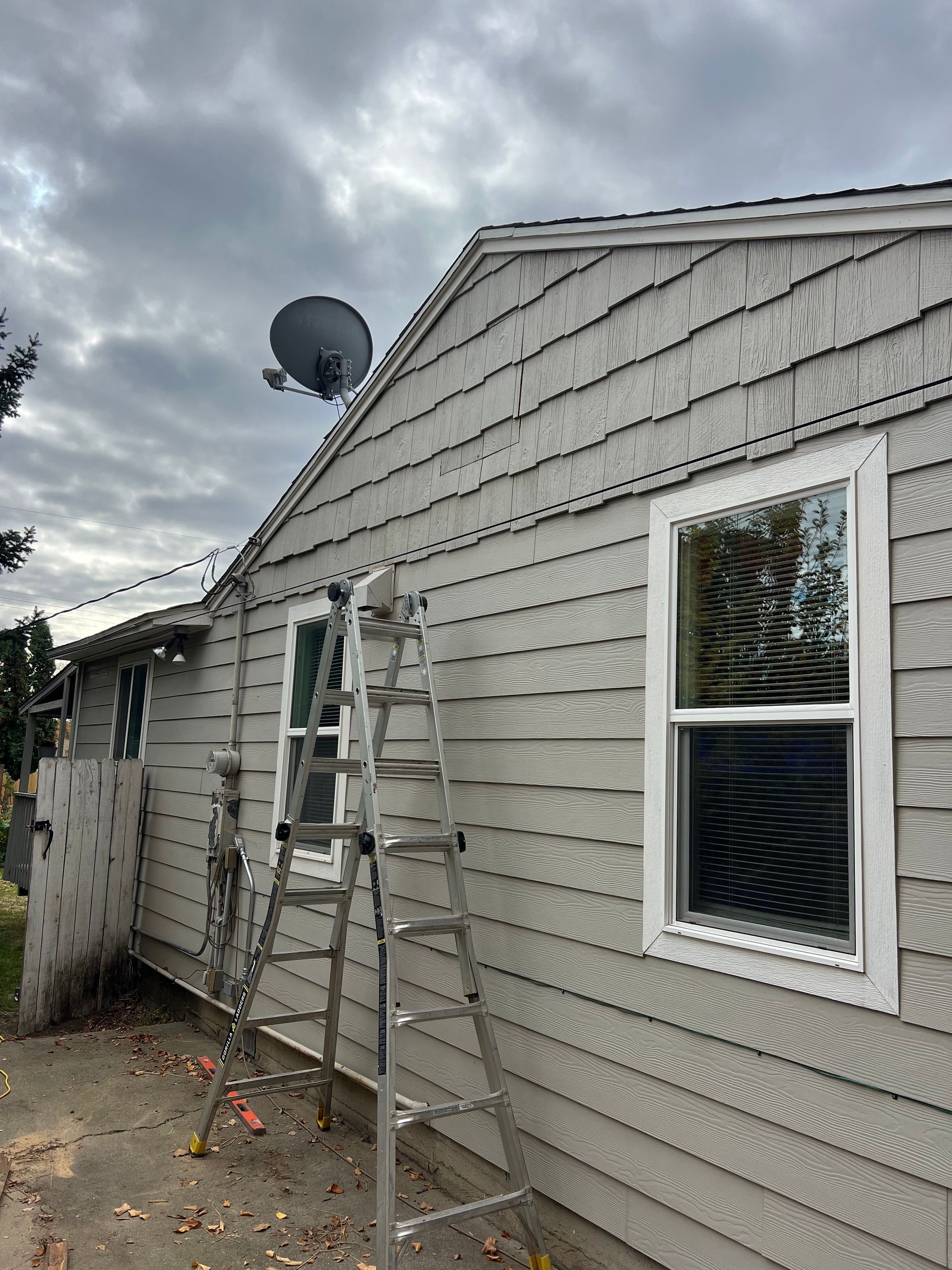 A gray-sided house with a satellite dish, windows, and a ladder against the wall under a cloudy sky.