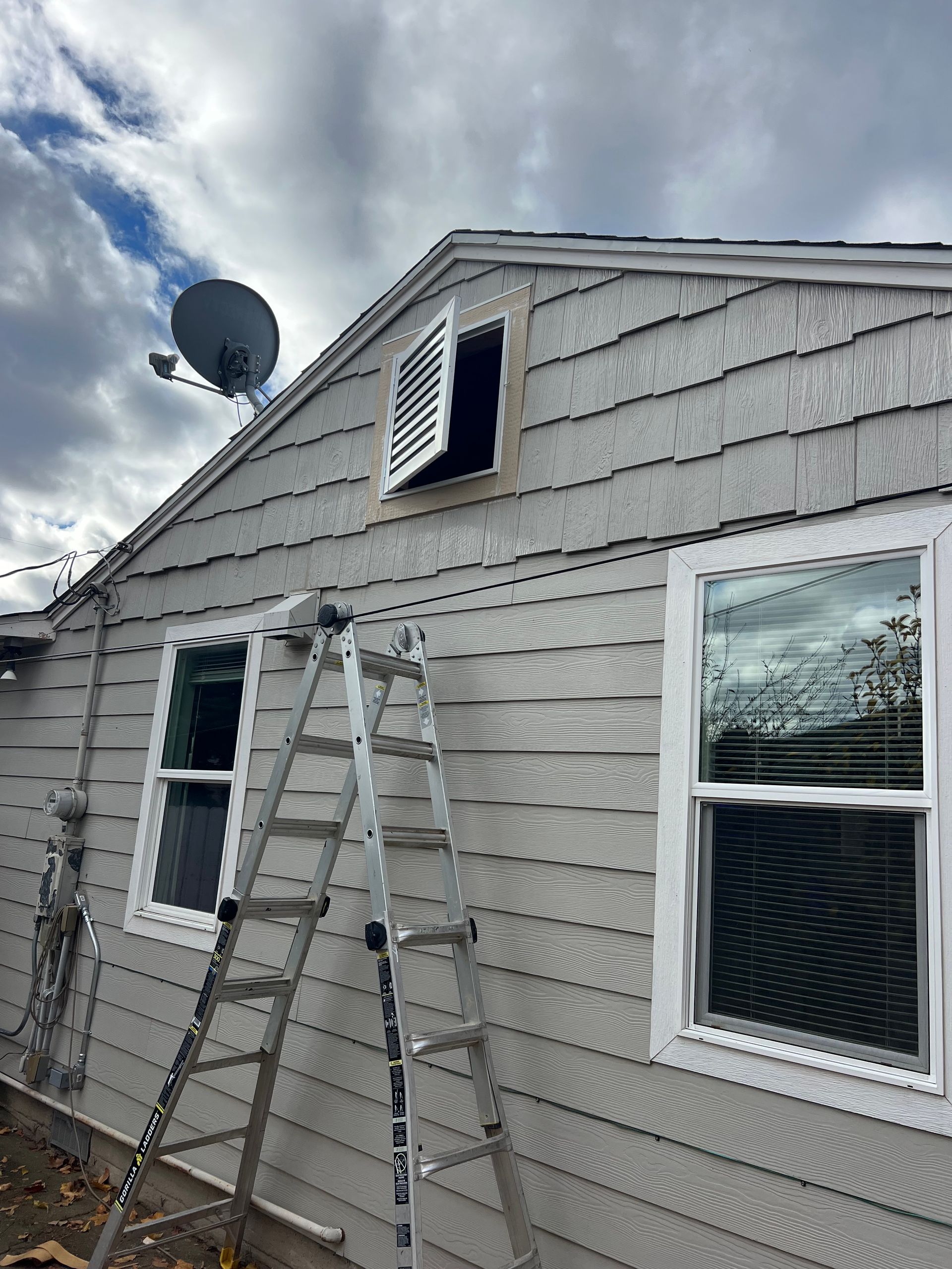 Exterior of a house with gray siding, an open attic vent, and a ladder propped against the wall.
