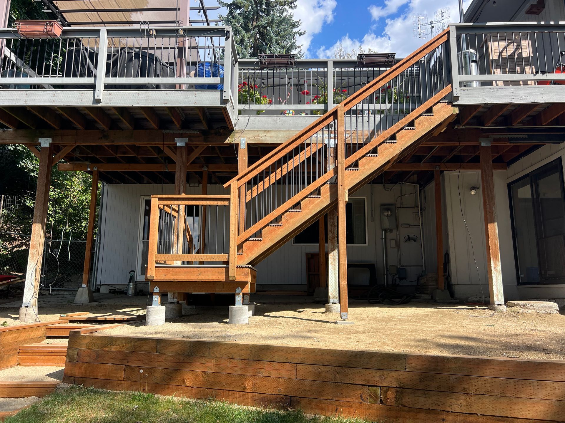 Two-story wooden deck with stairs. Brown wood railings and support beams. Light colored siding on a house.