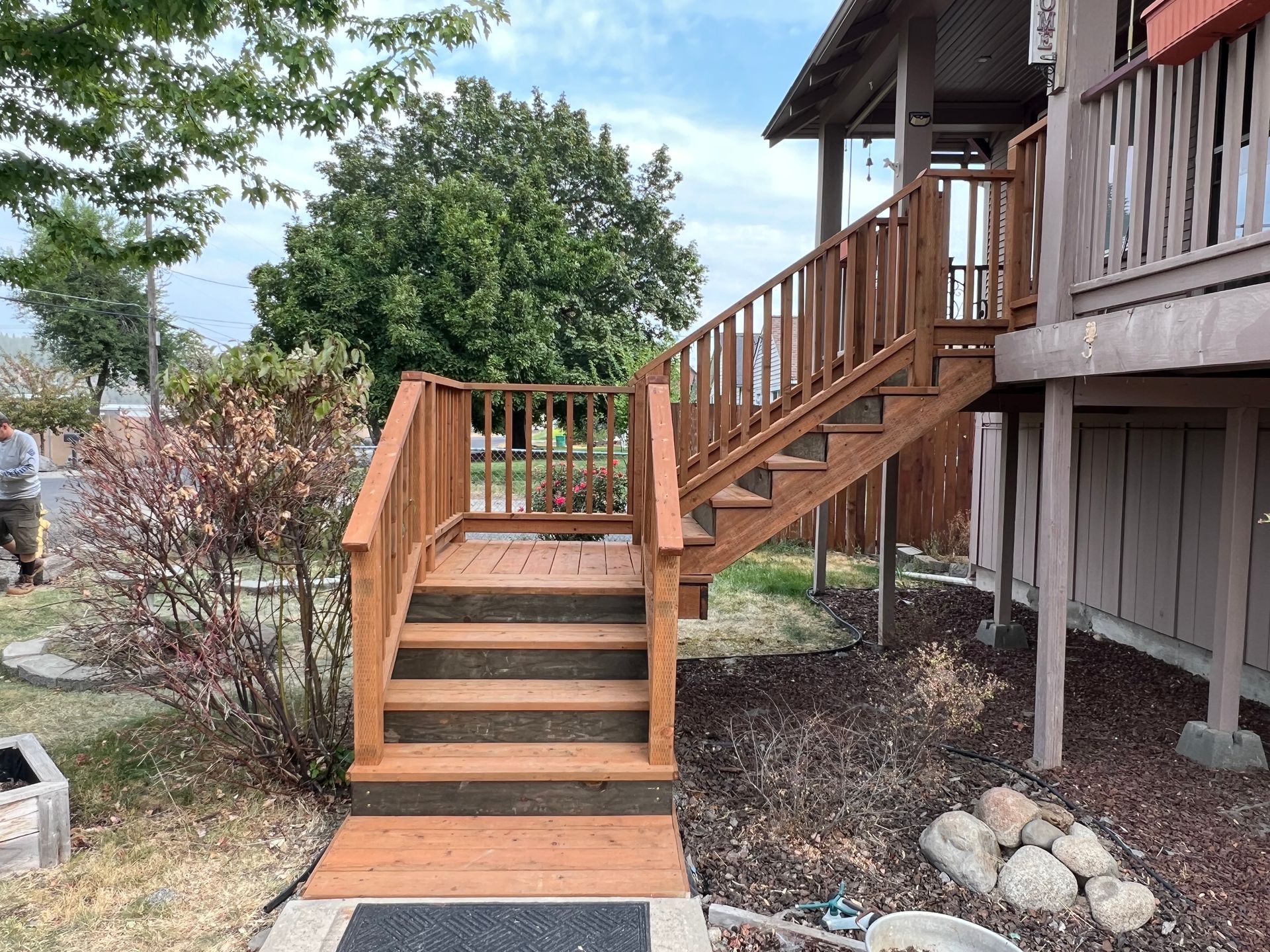 Wooden outdoor staircase leading to a porch. Brown stained steps and railings. Green bushes and a tree in the background.