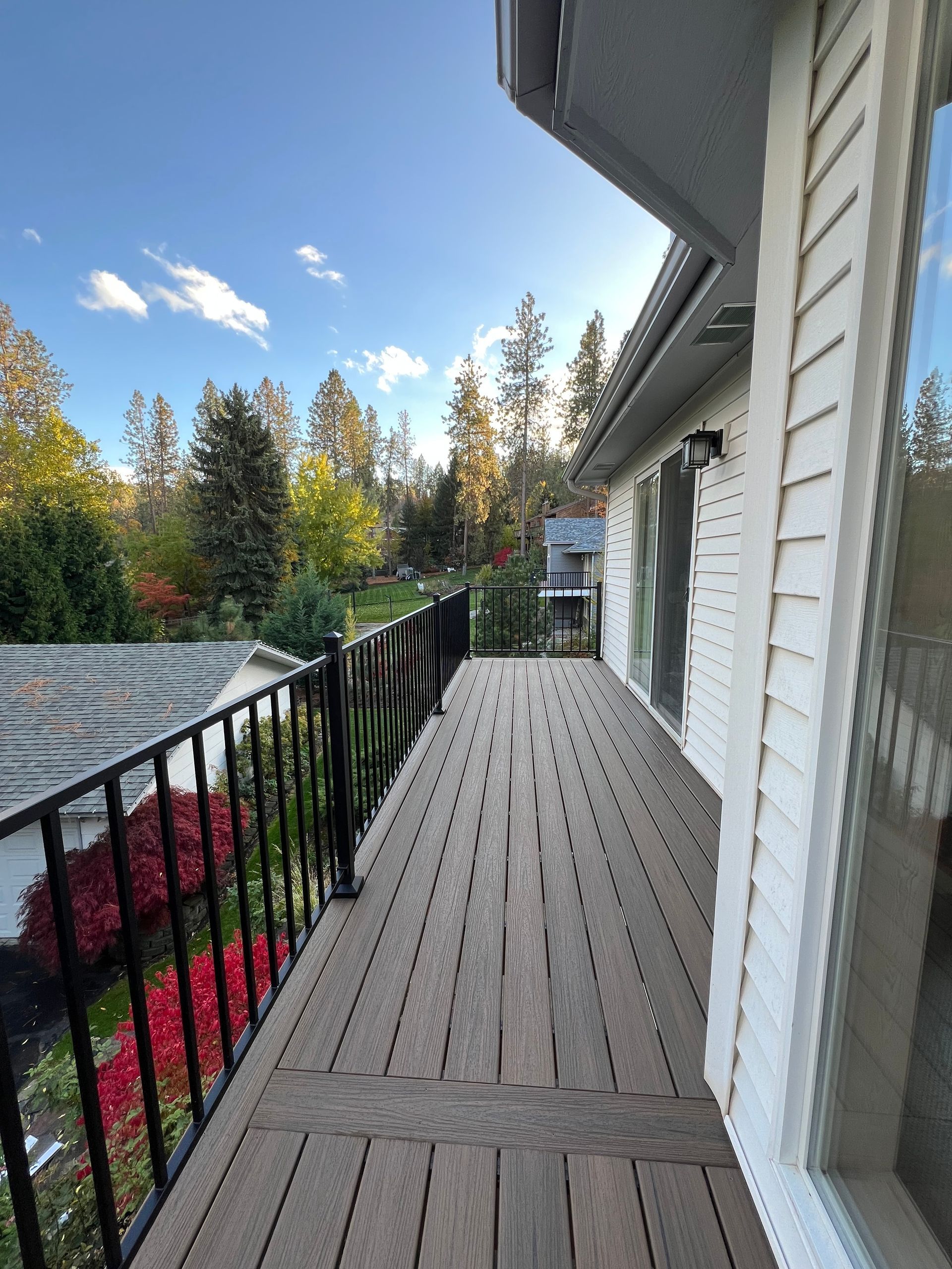 Wooden deck with black railing, white siding, and trees in the background under a blue sky.