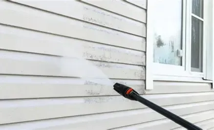 Power washer cleaning white siding on a house, next to a window.