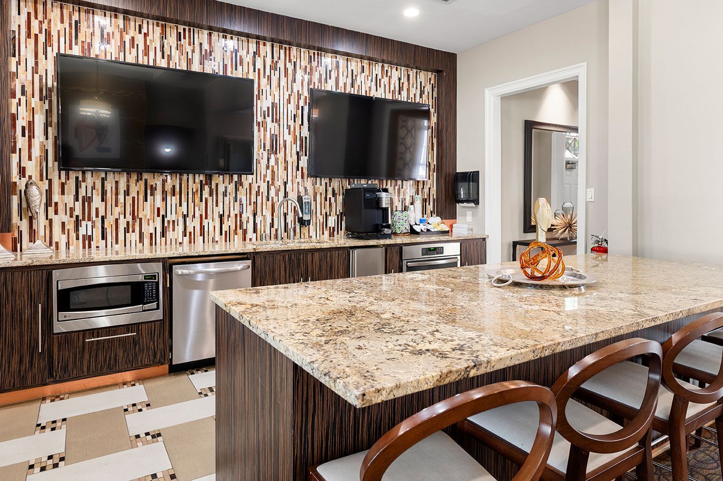 A communal kitchen area with granite counter seating, stainless steel appliances, two TVs, and a mosaic tile wall at Townhomes of Bent Tree, offers apartments in Dallas, TX.