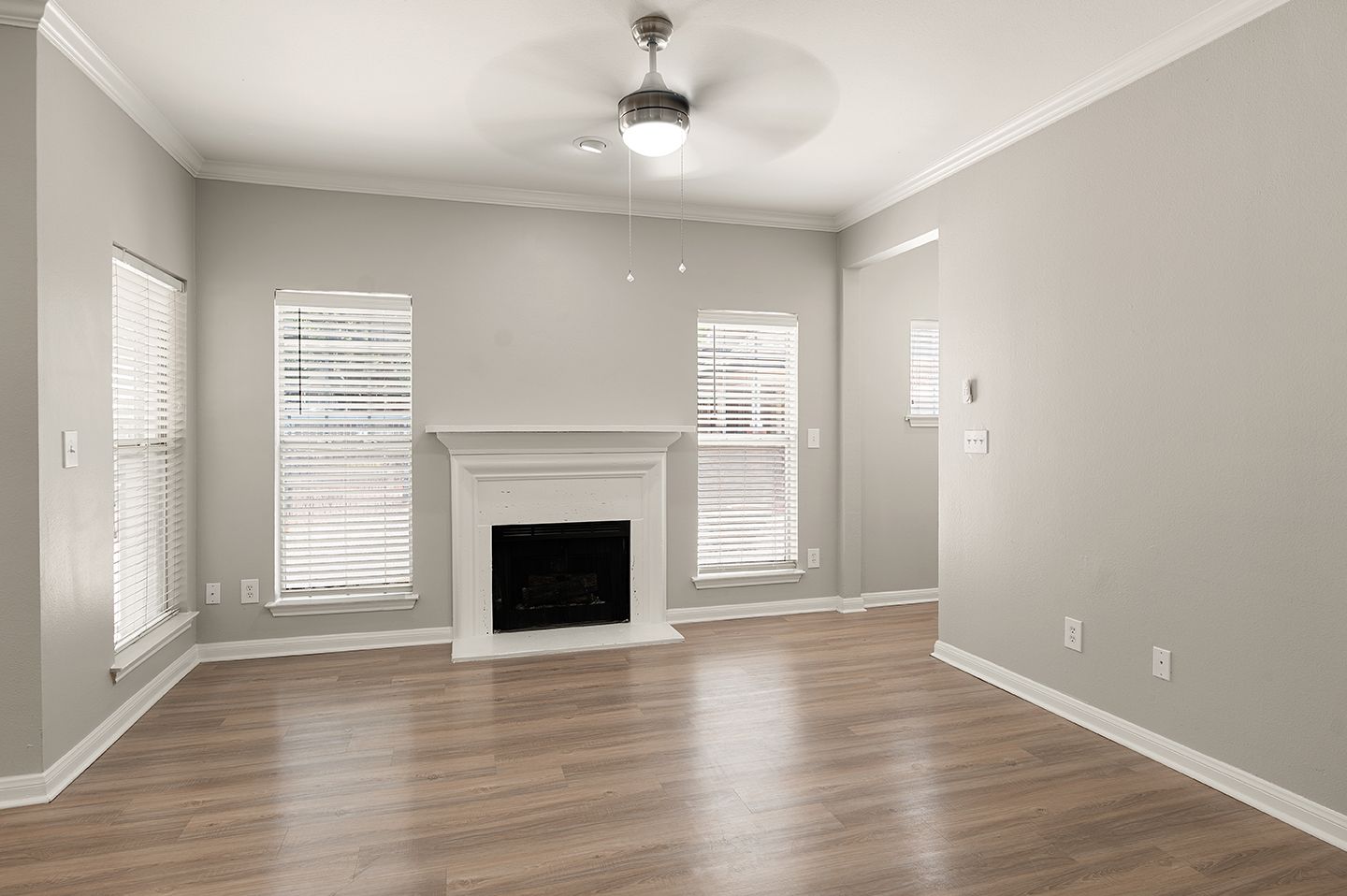 Empty living room with fireplace, three windows with blinds, hardwood floors, and a ceiling fan at Townhomes of Bent Tree, offers apartments in Far North Dallas, TX.