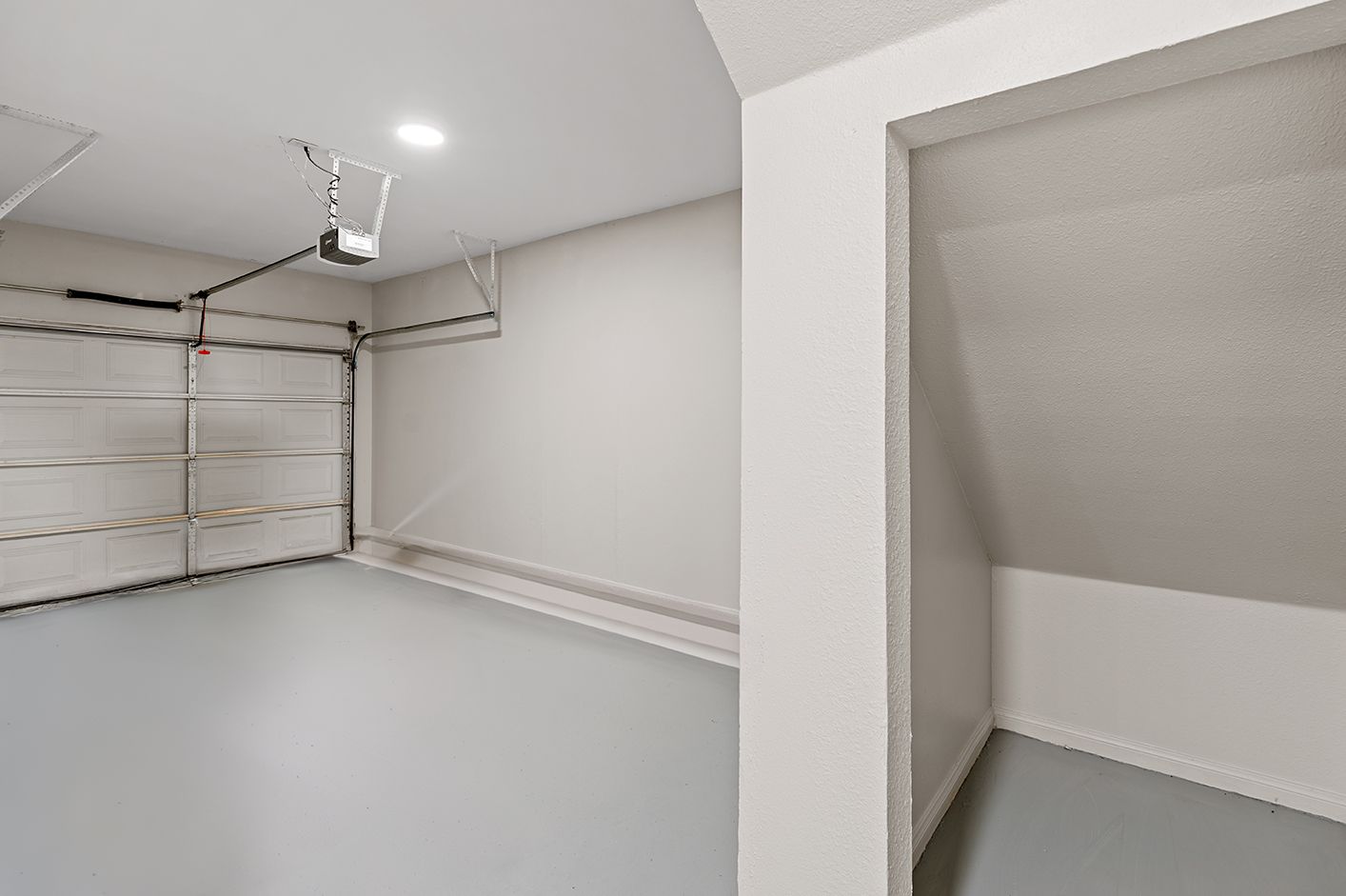 Empty garage with a closed garage door, white walls, and a gray floor. A storage nook is in the corner at Townhomes of Bent Tree, offers apartments in Far North Dallas, TX.