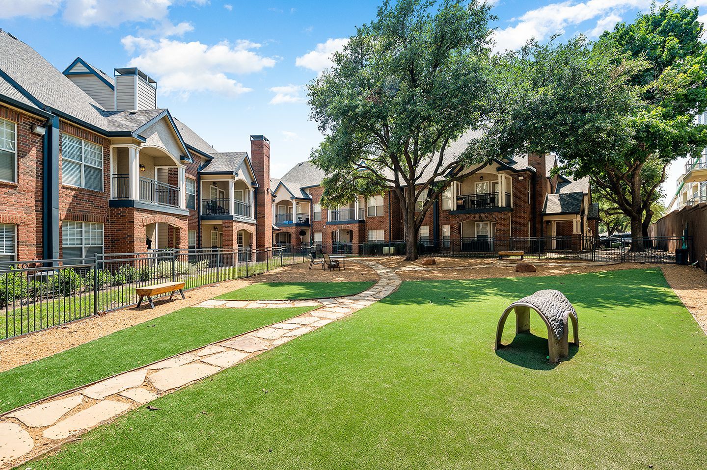 Apartment complex with green lawn and dog park, brick buildings, blue sky, and trees.