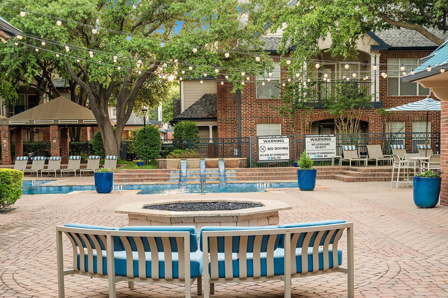 Courtyard with fire pit, pool, seating, and brick buildings. Warm colors and string lights at Townhomes of Bent Tree, offers North Dallas apartments.