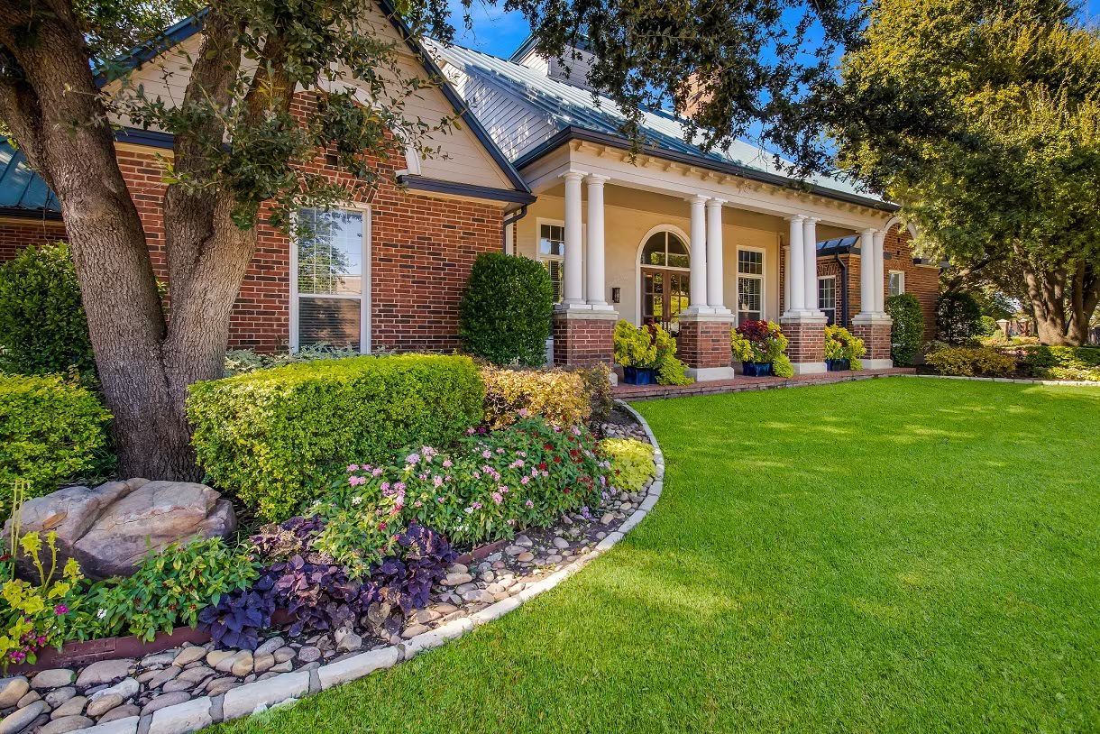 House with a porch, brick exterior, and well-manicured lawn and landscaping, under a bright blue sky at Townhomes of Bent Tree, offers Far North Dallas apartments for rent with garages.