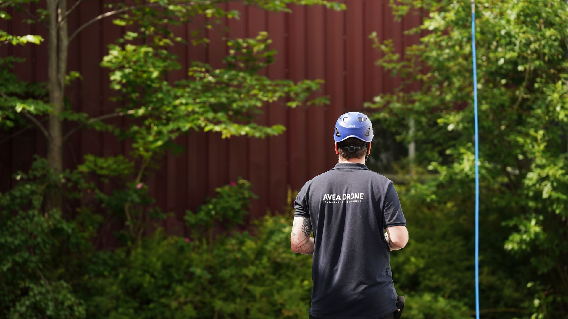 En mann med blå hjelm og svart skjorte står foran et rødt hus med drone til vedlikehold med fasadevask.