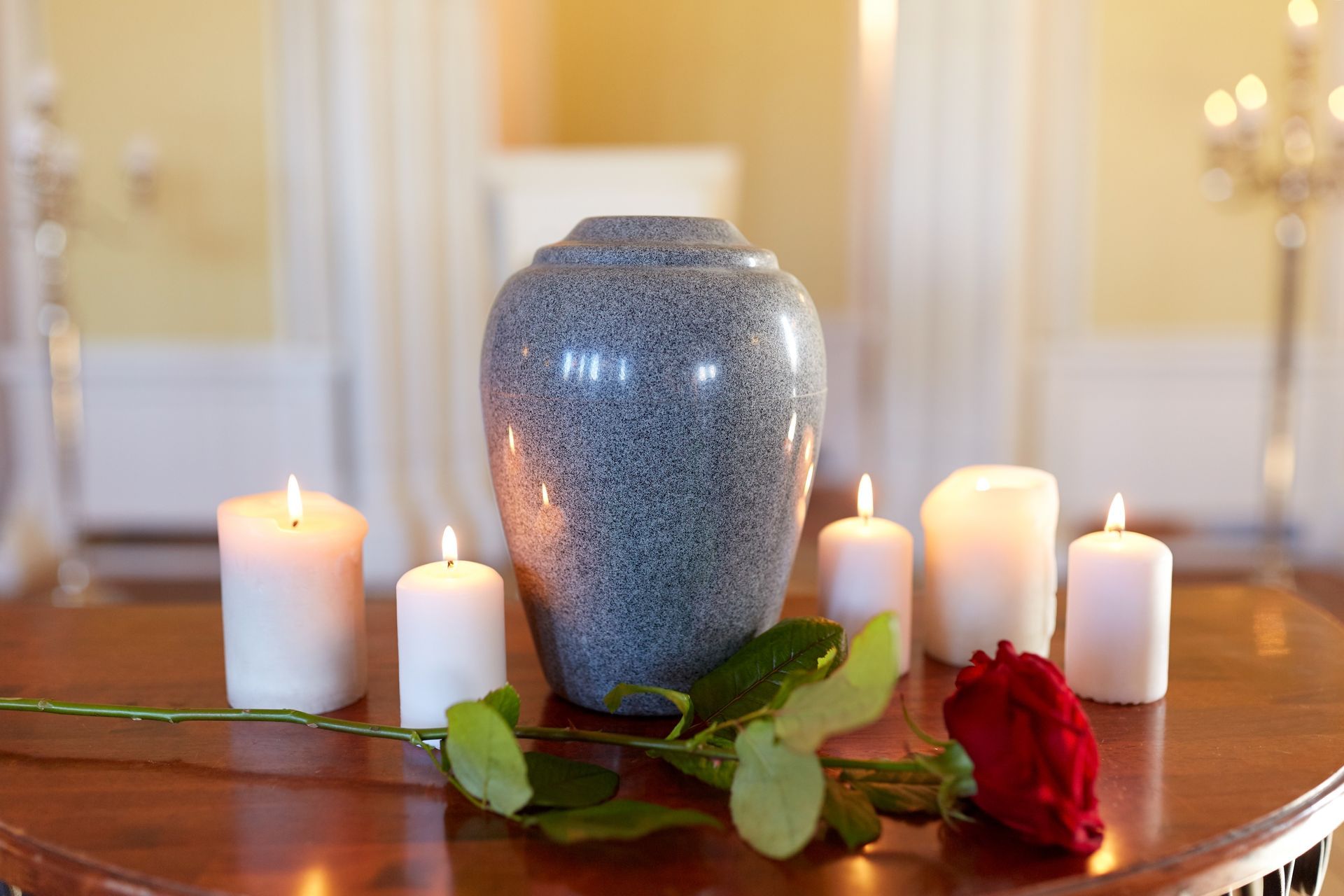 A gray urn with lit candles and a red rose on a wooden table in a room.