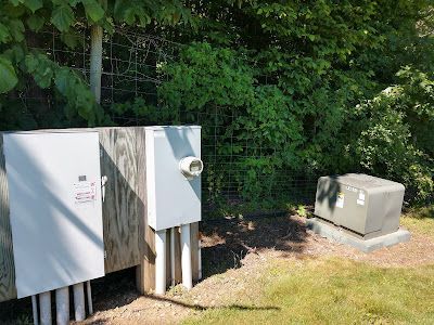 Outdoor electrical boxes beside a fence and foliage on grass.