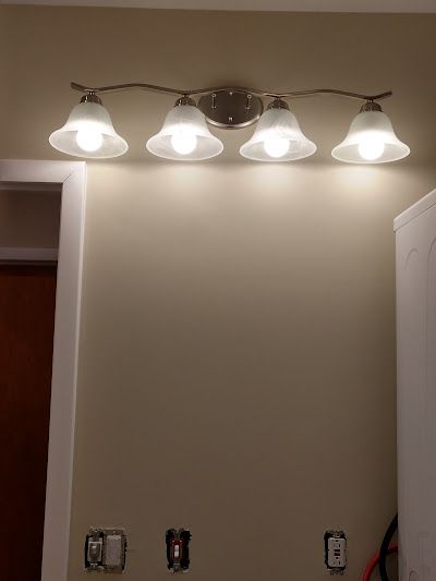 Four-bulb vanity light fixture mounted above unfinished wall. Beige wall, white lights, and light fixture is brushed metal.