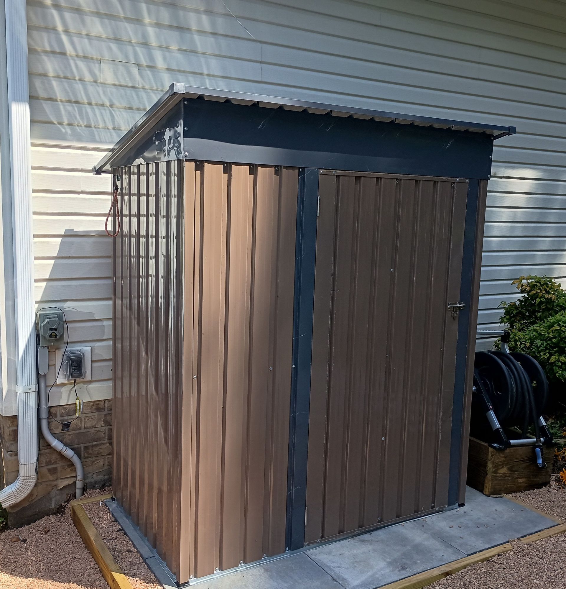 Brown metal storage shed against a light-colored wall, with a corrugated metal roof and a concrete pad.