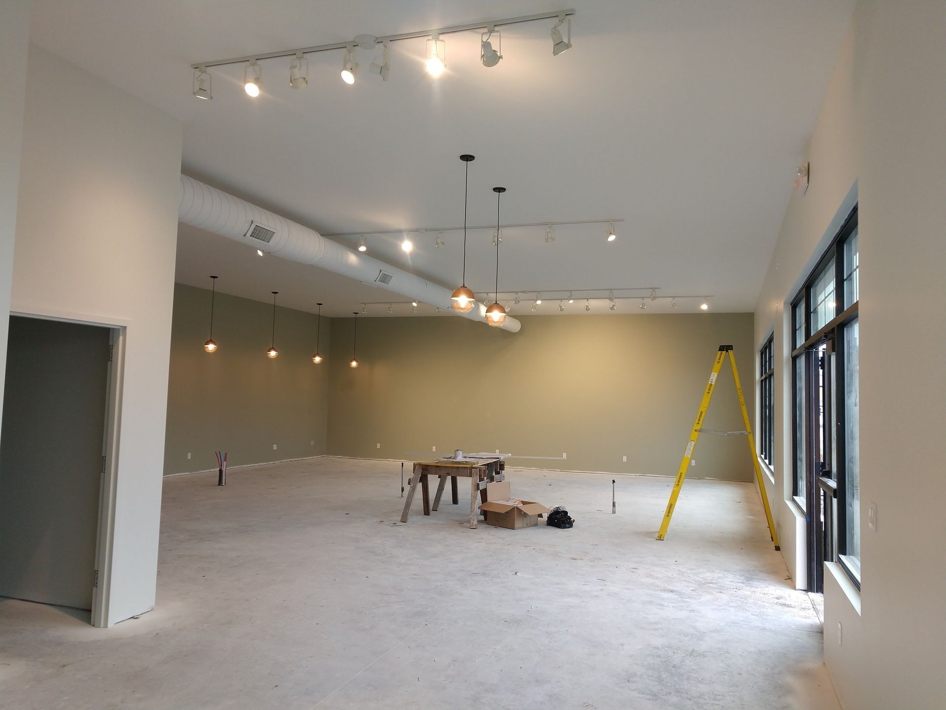 Empty commercial space with construction materials and lighting fixtures; a ladder leans against the wall.