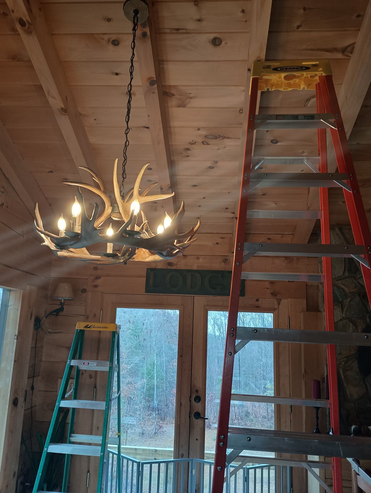 Antler chandelier hanging from a wood ceiling, with two ladders set up inside a cabin.